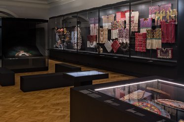 A view of an exhibition space featuring display cases with textiles and artifacts. The walls showcase colorful fabrics and patterned items. In the foreground, there are modern display tables and a darkened section with a large artifact, highlighting the artistic heritage on display.