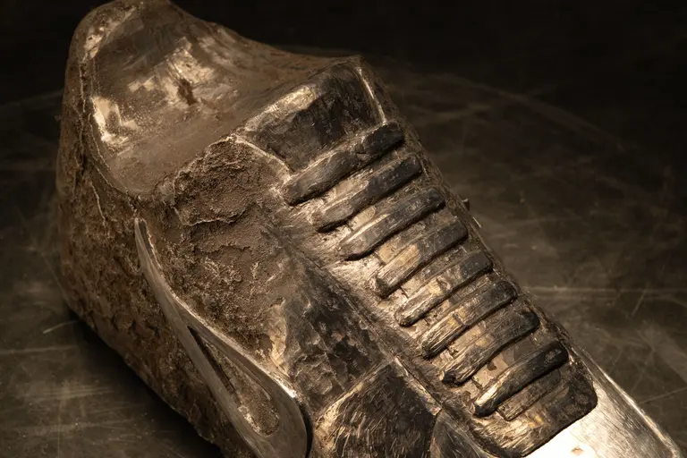 A close-up view of a worn sneaker upper, featuring visible scuff marks and a weathered texture. The shoe has a textured lace area and a portion that appears to be made of a metallic material. The background is dark, highlighting the shoe's details.