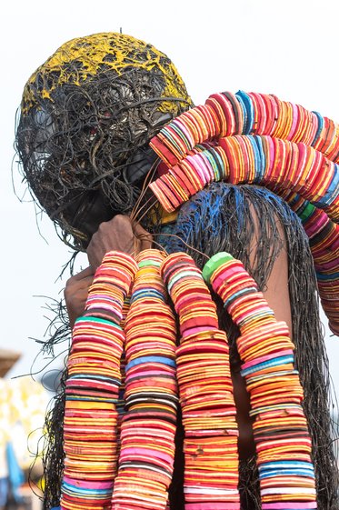 Eine Person mit einem bunten, spiralförmigen Kragen aus runden, bunten Scheiben trägt einen Kopf mit schwarzem, geflochtenem Material. Die Szene zeigt das Interesse an traditioneller Kleidung oder Veranstaltungen, die bunte Accessoires verwenden.