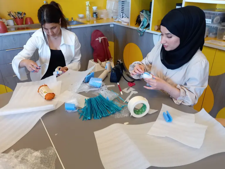 Zwei Frauen sitzen an einem Tisch, umgeben von verschiedenen Bastelmaterialien. Sie schneiden und gestalten Objekte. Die Umgebung ist kreativ gestaltet, mit bunten Akzenten. Beide Frauen konzentrieren sich auf ihre Arbeiten und zeigen handwerkliches Geschick.