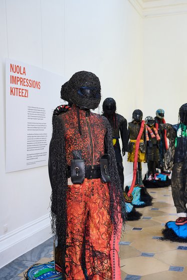 Eine Kunstinstallation zeigt mehrere skulpturale Figuren, die mit schwarzem Faden und bunten Materialien bedeckt sind. Eine Figur im Vordergrund trägt einen Helm und auffällige Kleidung in Rot und Schwarz. Im Hintergrund sind weitere Figuren zu sehen, die in unterschiedlichen Positionen stehen.