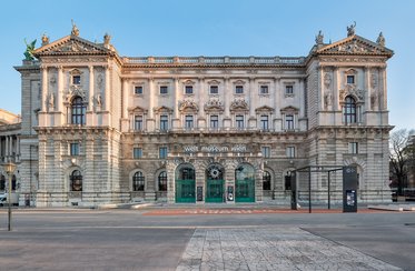 Das Bild zeigt die beeindruckende Fassade des Welt Museum Wien. Das historische Gebäude ist reich verziert mit Statuen und architektonischen Details. Die Eingänge sind durch große Fenster gekennzeichnet. Das Museum ist von einem klaren blauen Himmel umgeben und liegt in einer weitläufigen, ruhigen Umgebung.