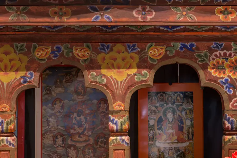 Das Bild zeigt einen kunstvoll verzierten Holzrahmen mit farbenfrohen floralen Mustern. Im Hintergrund sind religiöse Bilder sichtbar, die Teil eines kulturellen Erbes sind. Die Kombination aus Ornamenten und Bildern vermittelt eine spirituelle Atmosphäre.