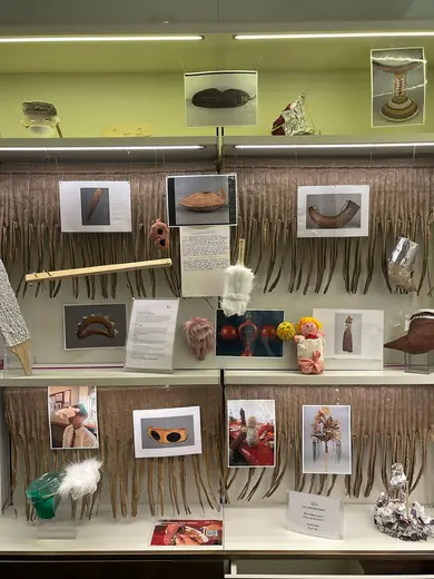 A display featuring various cultural artifacts, including masks, utensils, and sculptural pieces. Several photographs of these items are arranged on a backdrop of hanging strands. Descriptive texts are placed nearby, providing context for the artifacts. The overall layout is organized on light-colored shelves.