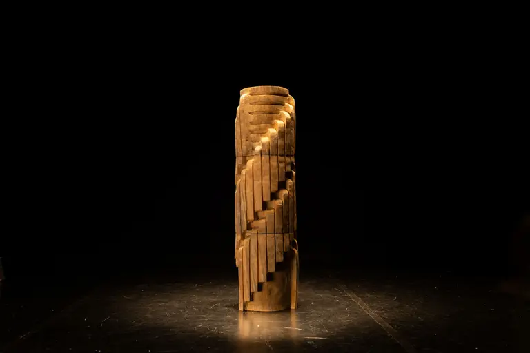 A tall, cylindrical wooden sculpture with intricate, spiral stair-like carvings. The piece is illuminated against a dark background, highlighting its texture and design.