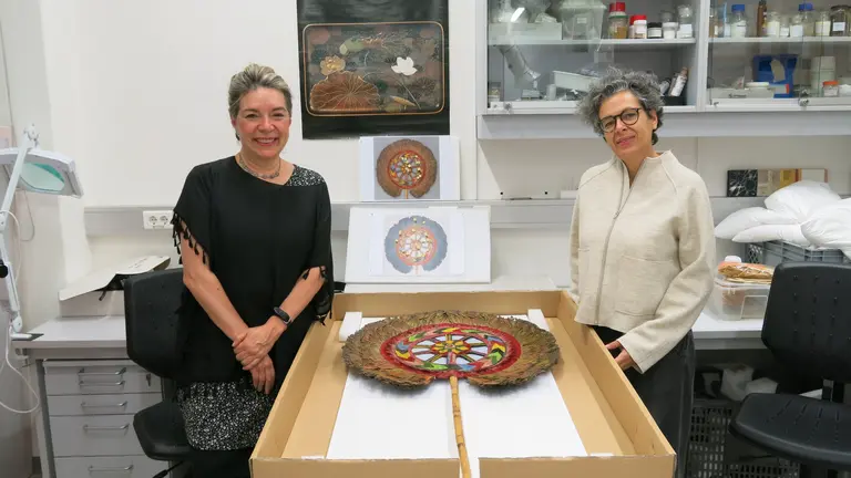 Zwei Frauen stehen neben einem Tisch, auf dem ein kunstvolles, farbenfrohes Objekt mit einem runden Muster liegt. Im Hintergrund sind Fotografien des gleichen Objekts sichtbar. Die Umgebung wirkt wie ein Arbeitsraum mit Regalen voller Laborbedarf.