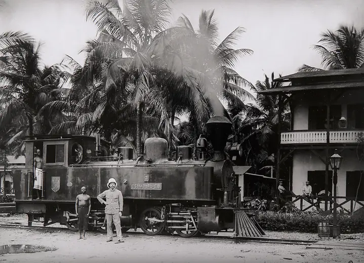 A historical black-and-white photograph features a steam locomotive parked near a building surrounded by palm trees. Two men, one in a uniform and the other in local attire, stand beside the train, which has a prominent logo displayed. The setting suggests a tropical environment.
