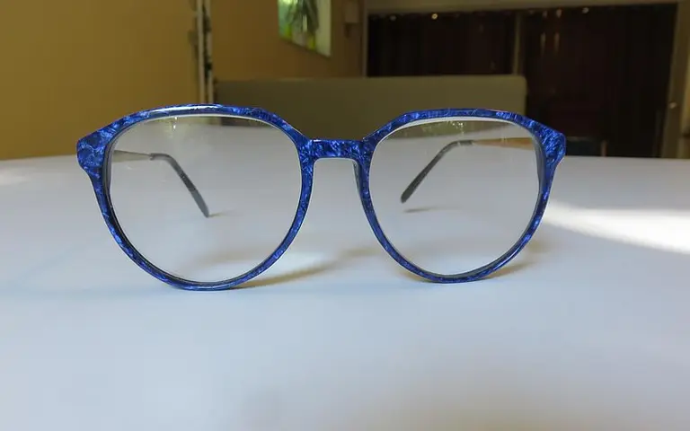 Ein Paar blauer Brillen mit runden Gläsern liegt auf einem weißen Tisch. Die Fassung ist auffällig und hat ein strukturiertes Muster. Die Gläser sind klar und transparent, ohne Sichtbare Stärke.