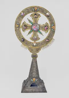 A decorative monstrance featuring an ornate cross shape with a circular frame. The frame is embellished with colorful gemstones and intricate golden details, highlighting a central glass section. The base has a detailed pattern, adding to the overall artistic design.