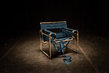 Ein moderner Stuhl mit einem metallischen Rahmen, der mit blauen Bändern umwickelt ist. Die Bänder hängen teilweise herab und verleihen dem Stuhl ein kreatives, künstlerisches Aussehen. Der Hintergrund ist dunkel, wodurch der Stuhl hervorgehoben wird.