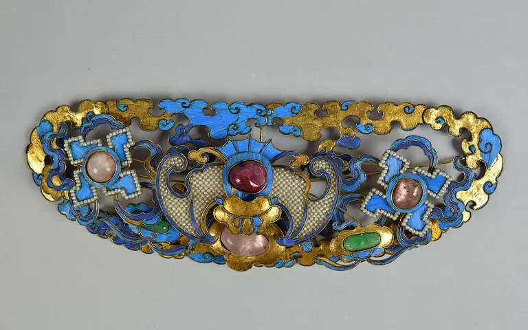 An ornate decorative piece featuring intricate designs in blue, gold, and various gemstones. The composition includes swirling patterns and shapes resembling crosses, punctuated by pink, red, and green stones, showcasing a blend of craftsmanship and color.