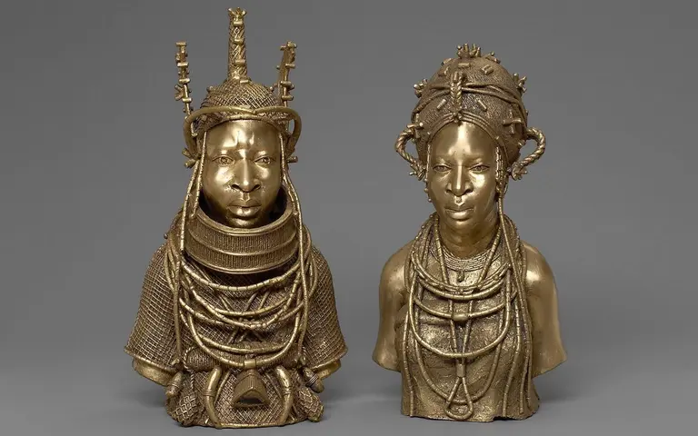 Zwei bronzene Skulpturen von menschlichen Köpfen, die traditionelle afrikanische Kleidung und Schmuck tragen. Die linke Figur hat eine komplizierte Kopfdekoration, die rechte Figur trägt mehrere Halsketten. Beide zeigen detaillierte Gesichtszüge und sind auf einem grauen Hintergrund platziert.