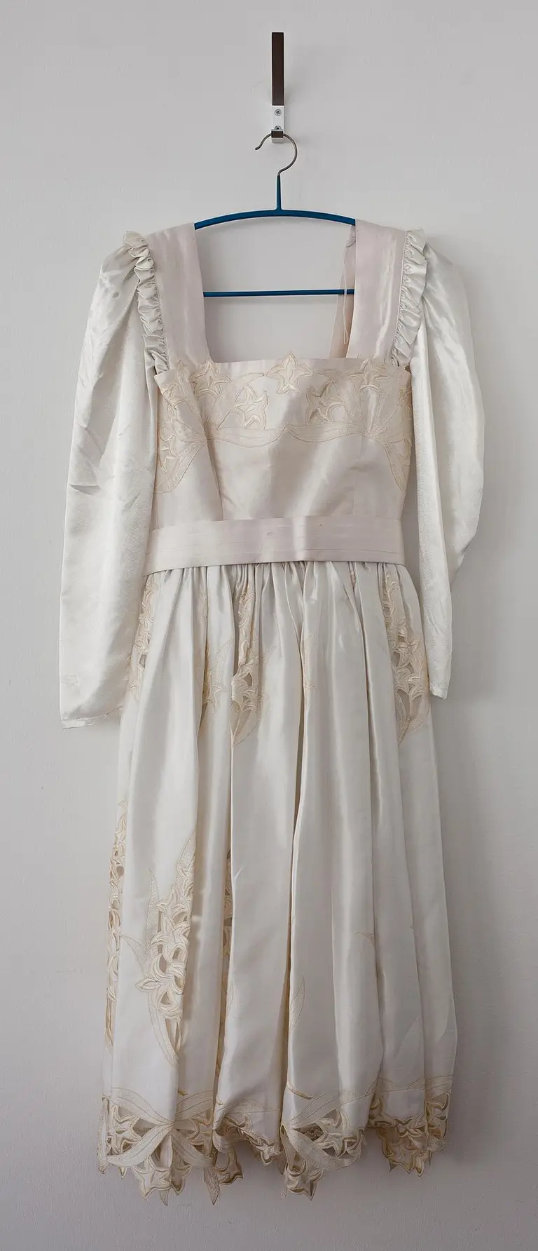 Ein traditionelles Kleid hängt an einem blauen Kleiderbügel. Es hat lange, rüschierte Ärmel und einen schlichten Oberteil mit feinen Verzierungen. Der Rock ist weit und endet mit einer dekorativen, brokatenartigen Spitze. Die Gesamtfarbe ist in sanften Weiß- und Cremetönen gehalten.