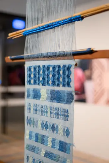 Ein Webstuhl mit einem hängenden Webstück in verschiedenen Blau- und Grüntönen. Das Gewebe zeigt unterschiedliche Muster und Texturen, die kunstvoll in sorgfältiger Handarbeit erstellt wurden.