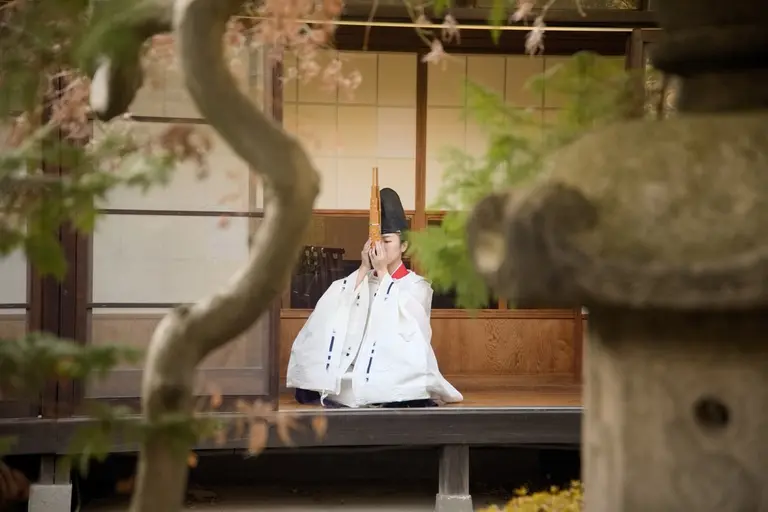A person in traditional attire kneels in a serene outdoor garden setting, holding a ritual implement. The background features a wooden structure with sliding doors and greenery. A stone lantern is partially visible, adding to the tranquil atmosphere.