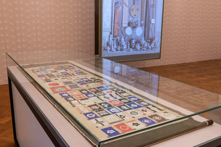 A glass display case contains a textile featuring various symbols and designs in colors such as blue, red, and white. In the background, a large screen shows a collection of antique objects, enhancing the educational context of the exhibit.