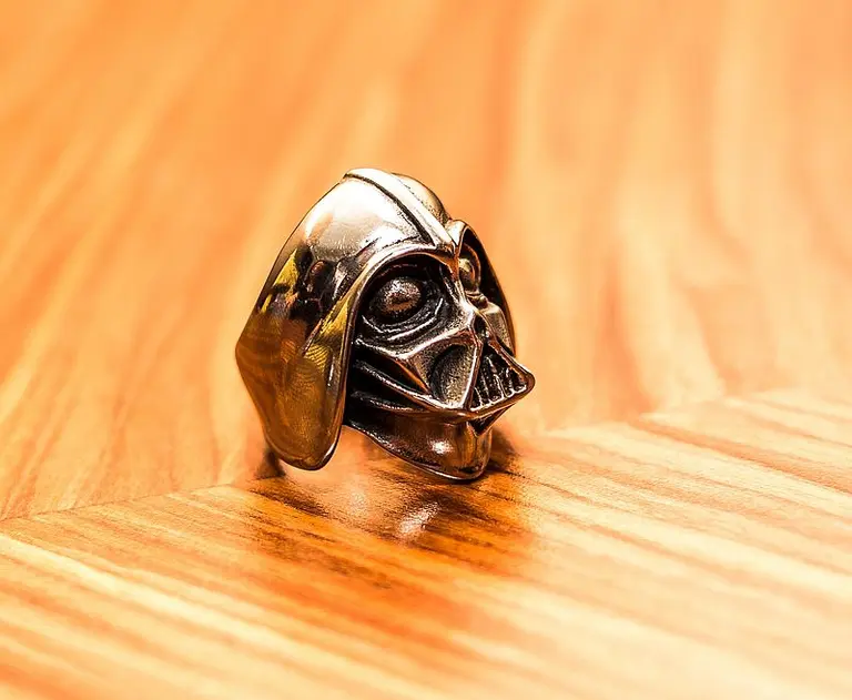 Ein silberner Ring in Form eines Darth Vader-Kopfes liegt auf einer Holzoberfläche. Der Ring zeigt detailreiche Züge des Charakters, einschließlich der ikonischen Helmform und des Gesichts.