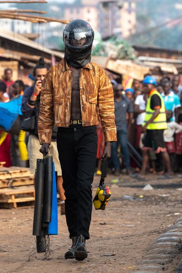 Ein Mann mit einem Helm und einer leicht glänzenden Jacke geht in einer belebten Straße. Er trägt mehrere Rohre in der einen Hand und ein weiteres Gegenstand in der anderen Hand. Im Hintergrund sind Menschen und Marktstände zu sehen.