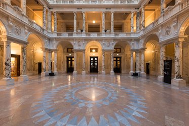A spacious, elegant interior of a historic building features intricate marble columns and a patterned floor design. Natural light illuminates the area, highlighting the ornate architecture and the arched openings leading to other rooms.
