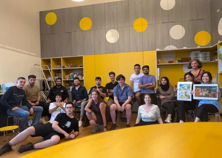 Eine Gruppe von jungen Menschen sitzt und steht in einem hellen Raum mit gelben und grauen Wänden. Sie lächeln und posieren, einige halten Bilder in den Händen. Die Atmosphäre wirkt freundlich und gemeinschaftlich.