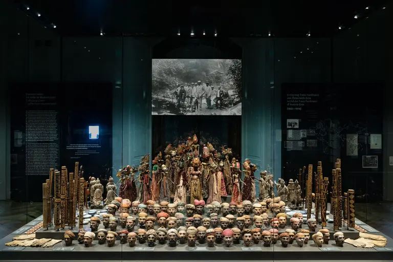 Eine Ausstellung zeigt eine Ansammlung von Totenschädeln und kunstvoll gestalteten Figuren. Im Hintergrund ist ein historisches Foto von Menschen zu sehen. Die Objekte sind auf einem glasernen Tisch angeordnet, umgeben von dunkel gehaltenen Wänden mit Informationshinweisen.