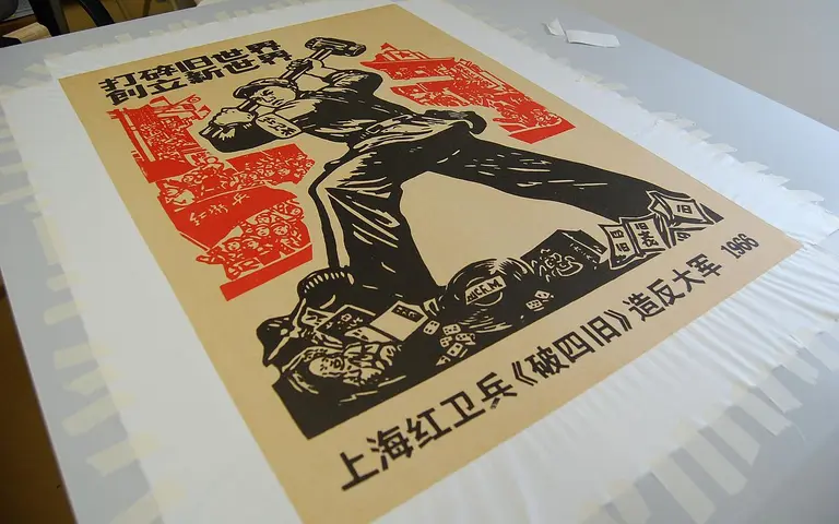A vintage propaganda poster featuring a strong figure holding a hammer, surrounded by red and black illustrations. The artwork emphasizes themes of labor and industry, with text in Chinese at the bottom. The poster is partially adhered to a surface with masking tape around the edges.