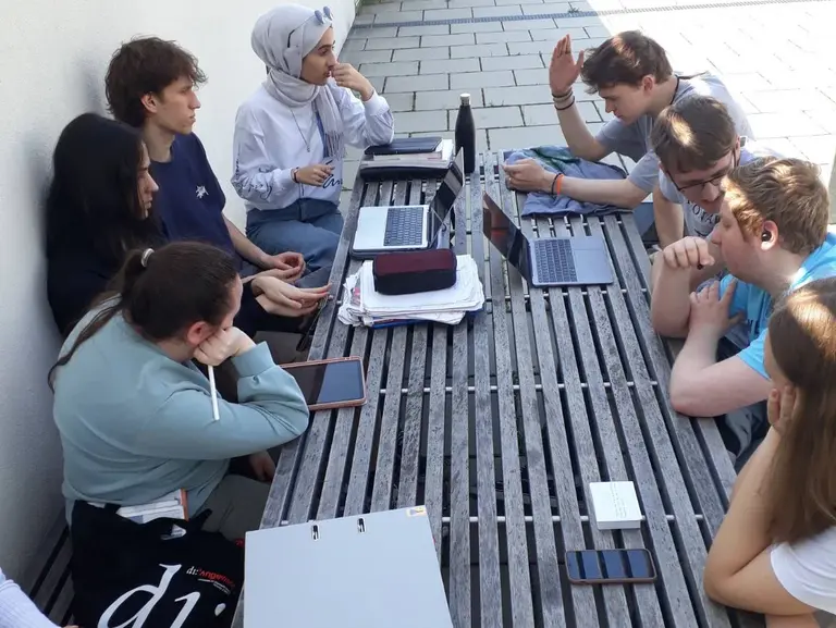 Eine Gruppe von zehn Personen sitzt an einem Tisch im Freien. Einige haben Laptops und Tablets vor sich. Sie scheinen an einer Diskussion oder einem Projekt zu arbeiten. Der Hintergrund zeigt einen klaren Himmel und eine ruhige Umgebung.