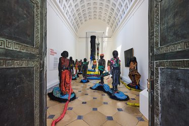 Blick durch kunstvolle Türen in einen Ausstellungsraum, in dem eine Gruppe von farbenfrohen Skulpturen in menschlicher Form steht. Die Figuren sind auf einer dekorativen Bodenfläche verteilt, während im Hintergrund ein größerer, zentraler Kunstgegenstand sichtbar ist.