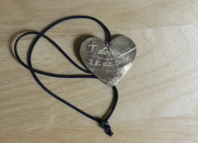 Das Bild zeigt ein herzförmiges Medaillon aus Metall, das an einem schwarzen Band hängt. Auf der Vorderseite steht 