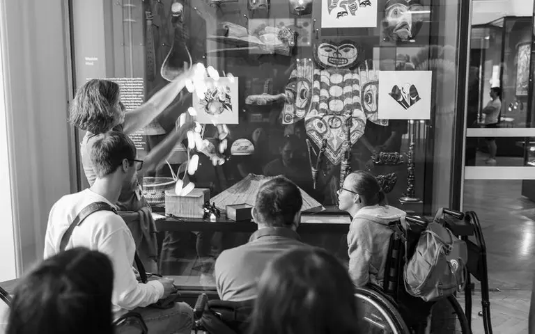 Eine Person zeigt Exponate in einem Museum, während eine Gruppe von Menschen, darunter zwei Personen im Rollstuhl, aufmerksam zuschaut. Im Hintergrund sind kunstvolle Gegenstände und Bilder zu sehen, die kulturelle Bedeutung haben.