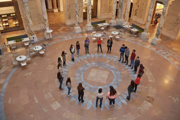Eine Gruppe von etwa 20 Personen steht in einem großen, runden Muster auf einem gefliesten Fußboden. Die Teilnehmer sind in einem Innenraum mit hoher Decke und Säulen angeordnet. Es gibt Tische und Stühle in der Umgebung, aber der Fokus liegt auf der Zusammensetzung der Gruppe.