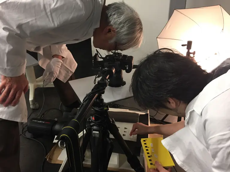 Two individuals in white lab coats are focused on a camera set on a tripod, examining a collection of small samples. One person is adjusting something or referring to a tool, while the other is peering through the camera viewfinder, indicating a scientific or research setting.