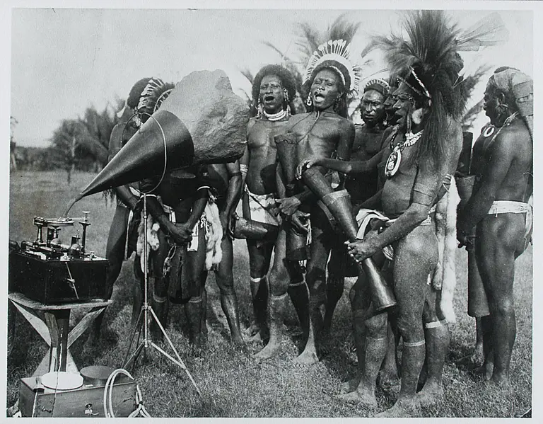 Eine historische Schwarz-Weiß-Aufnahme von mehreren indigenen Männer, die traditionell gekleidet sind und um ein Grammophon versammelt sind. Das Grammophon hat einen großen Trichter und steht auf einem Tisch. Im Hintergrund sind Palmen zu sehen.