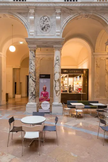 Ein gemütlicher Innenbereich mit Marmorsäulen und mehreren Tischen und Stühlen. Im Hintergrund ist ein Café sichtbar. Die Umgebung wirkt hell und einladend, ideal für einen entspannten Aufenthalt.
