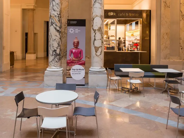 A spacious, elegantly designed indoor area features a circular table surrounded by chairs. In the background, a café with a counter and display can be seen, framed by tall marble columns and decorative arches. Soft lighting adds a warm atmosphere to the setting.