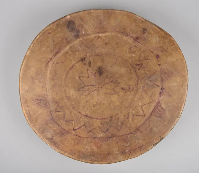 Eine runde, flache Platte aus einem hellen Material. Auf der Oberfläche sind geometrische Muster und eine zentrale Blütenform in dunklerer Farbe eingraviert. Die Platte hat eine unregelmäßige Form mit leicht erhöhten Rändern.