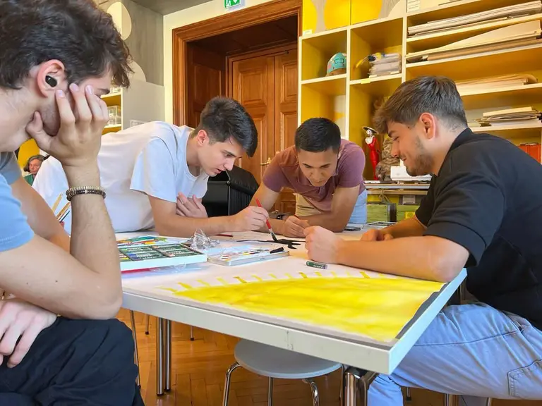 Vier junge Männer beugen sich über einen Tisch, auf dem Zeichenmaterialien liegen. Sie arbeiten gemeinsam an einem Projekt und scheinen fokussiert und kreativ zu sein. Der Hintergrund zeigt eine freundliche, helle Umgebung mit Regalen.