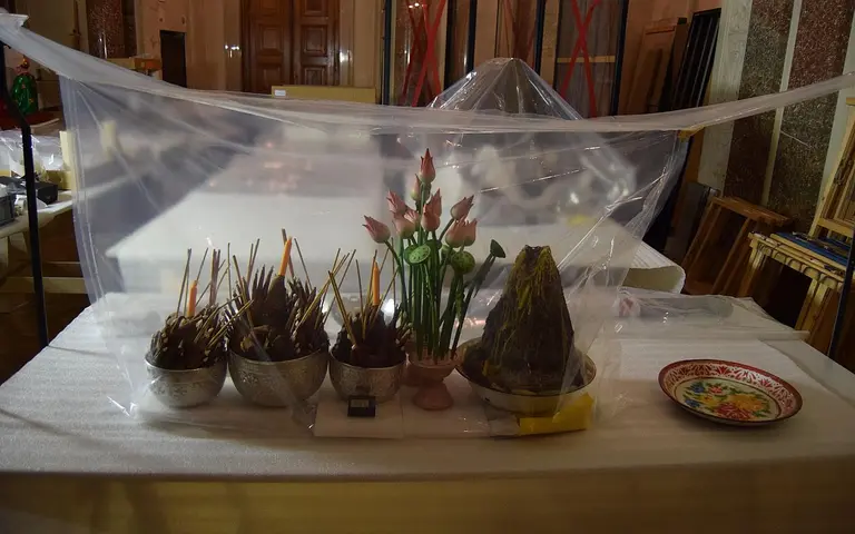 A covered display featuring multiple bowls with incense sticks, a small vase with artificial flowers, and a decorative item resembling a rocky formation. The setup is placed on a table, partially obscured by a translucent plastic sheet.