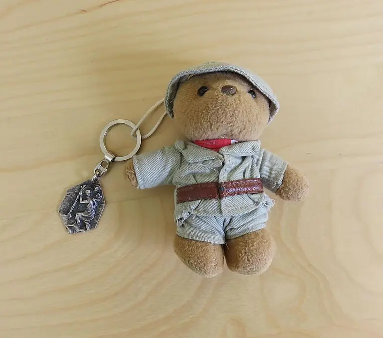 Ein kleiner Teddy mit einem Schlüsselanhänger, der einen hut und einen grauen Overalls trägt. Er hat ein rotes T-Shirt darunter und einen Gürtel um die Taille. Neben ihm hängt ein kleiner silberner Schlüsselanhänger mit einem Detail. Der Teddy liegt auf einer hellen Holzoberfläche.