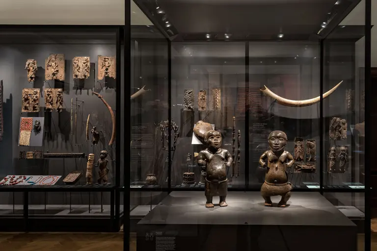 Das Bild zeigt eine Ausstellung mit verschiedenen kunstvollen Holzobjekten und Skulpturen. Im Vordergrund stehen zwei afrikanische Figuren, die geschichtlich bedeutend sind. Im Hintergrund sind weitere geschnitzte Kunstwerke und Artefakte in Vitrinen zu sehen, die kulturellen Reichtum repräsentieren.