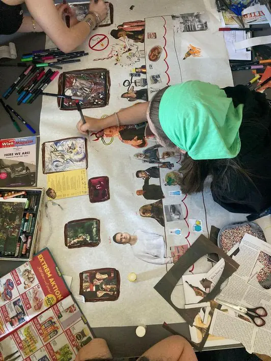 Eine Person mit einem grünen Kopftuch sitzt an einem Tisch und gestaltet eine kreative Collage. Sie zeichnet und platziert Bilder und Texte auf einem großen Blatt Papier, um ein visuelles Projekt zu erstellen. Rund um sie liegen verschiedene Stifte und Zeitschriften.