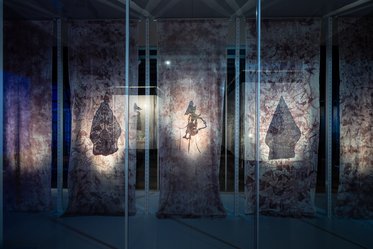 Alt text: A display featuring large, translucent fabric panels with abstract patterns. Backlit silhouettes of figures are visible on the panels, creating an artistic installation. The setting has dim lighting that enhances the visual contrast.