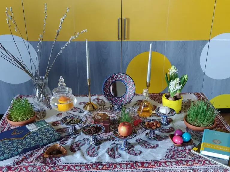 Auf dem Tisch liegt ein reich gedeckter Tisch mit traditionellen Gegenständen für das persische Neujahrsfest. Dazu gehören grüne Kräuter, Äpfel, Eier und Kerzen, umgeben von einer dekorativen Tischdecke. Im Hintergrund sind bunte Möbel zu sehen.