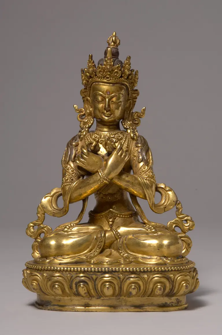 A golden statuette of a seated Buddha figure, adorned with intricate details. The figure has a serene expression, wears an elaborate crown, and holds its hands in a prayer position. The base features decorative patterns, highlighting the artistry of the sculpture.