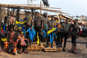 Eine Gruppe von Modellen steht auf Holzpaletten in einem belebten Markt. Sie tragen auffällige Kleidungsstücke mit künstlerischen Akzenten und Accessoires. Die Umgebung zeigt Holzkonstruktionen und eine Vielzahl von Menschen im Hintergrund.