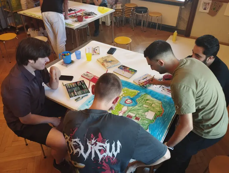 Vier junge Männer sitzen an einem Tisch, der mit Kunstmaterialien und Zeichnungen bedeckt ist. Sie diskutieren und arbeiten gemeinsam an einem kreativen Projekt. Der Raum ist hell und freundlich, mit Stühlen im Hintergrund. Einige Kunstwerke sind auf dem Tisch ausgebreitet.