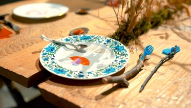 Ein gedeckter Tisch aus Holz mit einem bunten Teller, darauf liegt ein Löffel. Daneben liegen zwei weitere kleine Teller und verschiedene Utensilien, einschließlich farbiger Gabeln und Stäbchen. Die Tischoberfläche ist dekoriert mit Moos und kleinen Naturgegenständen.