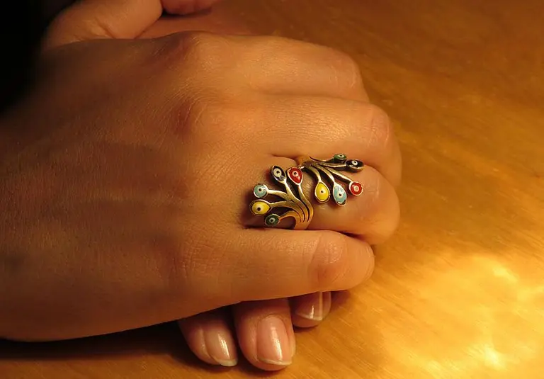 Eine Hand liegt auf einem Tisch, die oberste Fingerkuppe ist mit einem kunstvoll gestalteten Ring verziert. Der Ring hat ein organisches Design mit bunten Elementen in Blau, Rot und Gelb, das sich wie ein Blattwerk um den Finger windet.