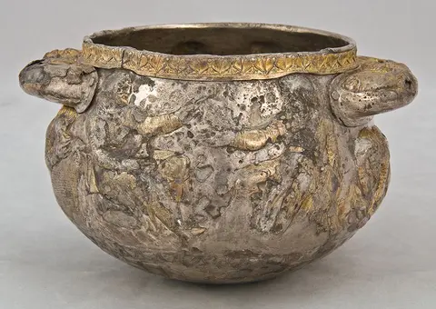 A decorative silver bowl with two handles, featuring intricate relief designs and a gold accent along the rim. The surface shows a textured pattern, indicating age and craftsmanship. The bowl has a rounded shape, typical of ancient vessels.
