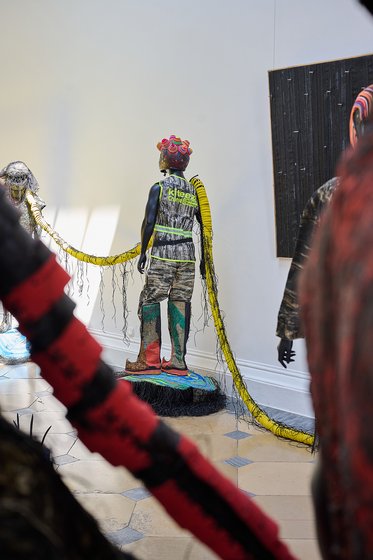 Eine künstlerische Installation zeigt eine figurative Statue mit bunten, auffälligen Details. Die Figur steht auf einem Podest und hat lebendige Accessoires. Im Hintergrund sind weitere Figuren und textile Elemente sichtbar, die eine dynamische Komposition im Raum schaffen.